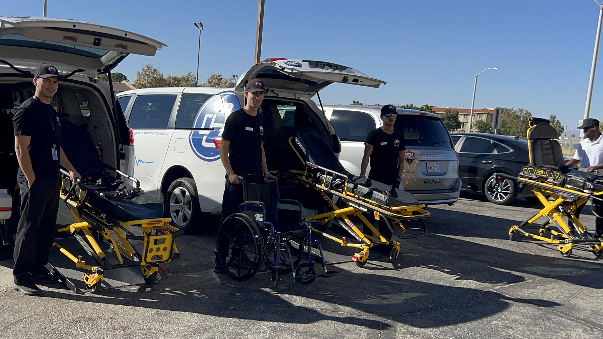 Crew members standing with gurneys, a wheelchair, and stair chairs behind an open transport van.